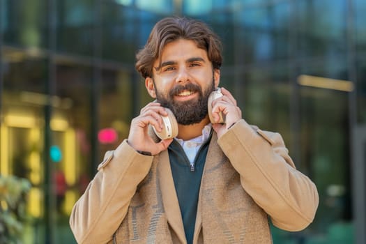 Joyful relaxed overjoyed Arabian business man listening favorite energetic disco rock n roll music outdoors. Adult guy taking off wireless headphones looking at camera smiling on downtown city street.