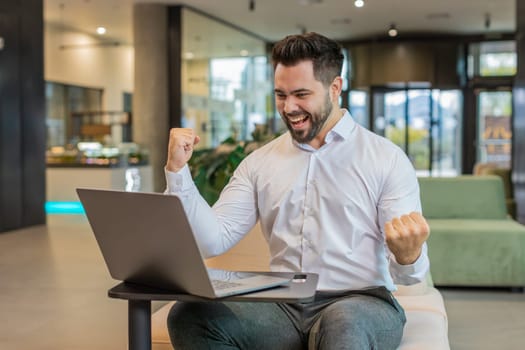 Caucasian bearded businessman working on laptop shocked by sudden victory, game winning lottery goal achievement, good career news. Excited guy freelancer clenching fists sitting in modern hotel lobby