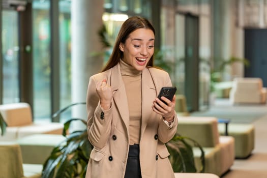 Happy amazed young businesswoman girl working on smartphone shocked by sudden victory game winning lottery jackpot achievement good career news. Excited woman in formal sitting in office hotel lobby.