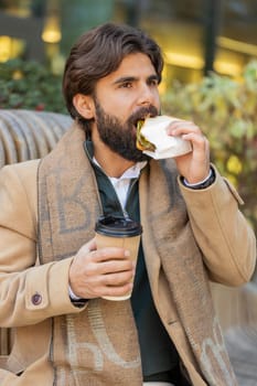 Indian young businessman tourist enjoy lunch break eating tasty sandwich meal and drinking hot beverage coffee or tea. Lebanese bearded man guy relaxing sitting on a bench on downtown city street