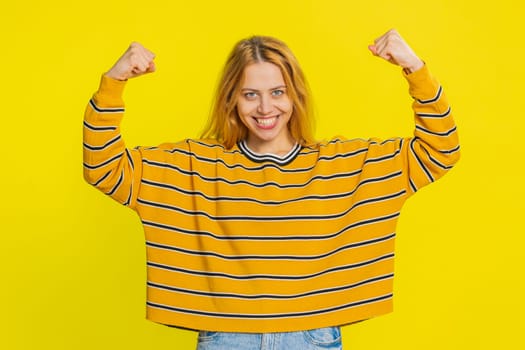 I am strong and independent. Happy smiling Caucasian woman showing biceps and looking confident, feeling power strength to fight for rights, energy to gain success win. Girl on yellow background