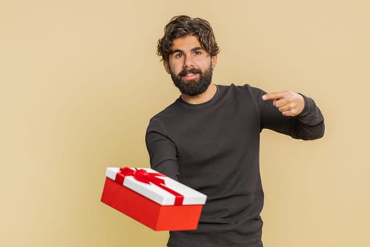 Positive smiling young Indian man presenting birthday gift box stretches out hands, offer wrapped present career bonus, celebrating party, sale. Bearded Hindu guy isolated on beige studio background.