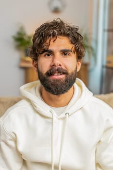 Portrait of smiling young Indian man looking at camera. Happy content adult bearded Hispanic guy in hoodie relaxing feeling satisfied concept sitting on sofa couch in living room at home. Lifestyles.