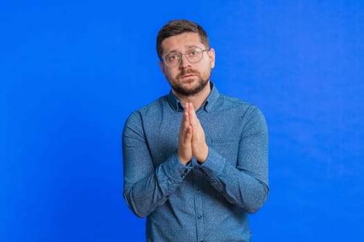 Please, God help. Caucasian middle-aged man praying, looking at camera and making wish, asking with hopeful imploring expression, begging apology, meditation. Adult guy isolated on blue background