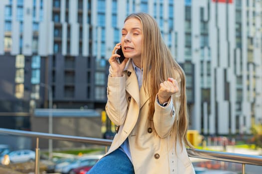 Angry mature businesswoman argues on smartphone while sitting on downtown city street. Dissatisfied middle-aged woman girl in coat shouts into the mobile phone, expressing frustration and a problem.