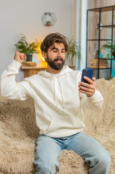 Happy young man using smartphone typing browsing saying yes, celebrating success victory, winning lottery jackpot goal achievement good positive news, triumph. Bearded guy at home sitting on sofa.