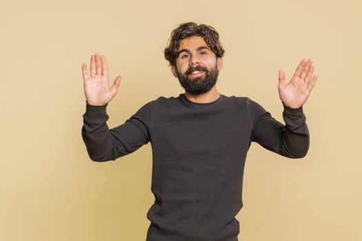Hello, welcome. Handsome young Indian man with long sleeves waving hi with his palm, greeting with hospitable friendly smile. Adult male Hispanic guy isolated on beige studio background indoors.