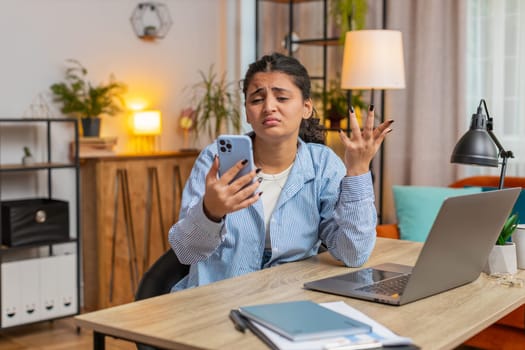 Upset displeased Indian woman freelancer using smartphone sitting at desk table in living room. Sad young girl touching head loses becoming surprised by sudden lottery results or bad news. Lifestyle.