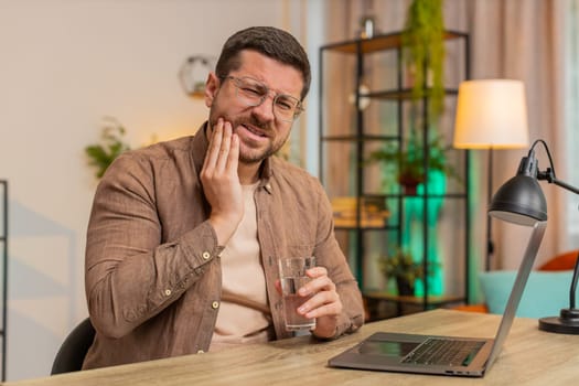 Caucasian young businessman freelancer sitting at home office table drinking cold water feeling toothache touching mouth, painful expression of dental illness, gum disease, dental disease concepts.