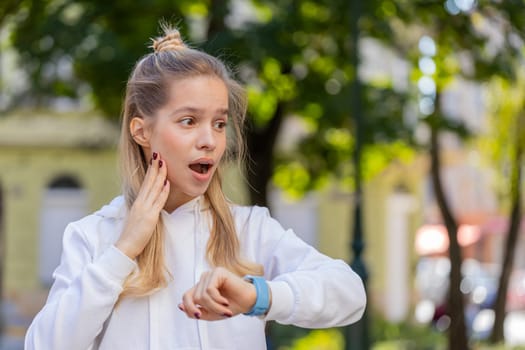 Young woman worrying to be punctual, with anxiety checking time on watch, running late to transport, being in delay, deadline outdoors. Girl teenager tourist walking in city street. Town lifestyles.