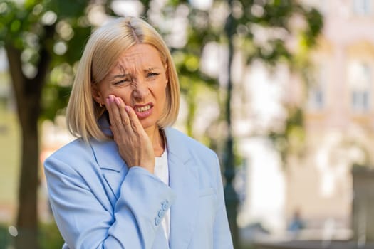 Mature Caucasian businesswoman in formal suit standing on city street feeling toothache bad unpleasant symptoms, suffering from pain holding hand on cheek jaw damaged dental nerve needs medical help.