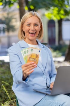 Happy mature businesswoman investor counting euros banknotes on city street. Smiling entrepreneur in suit looking at laptop holding money and rejoices at good earnings. Business success lottery.