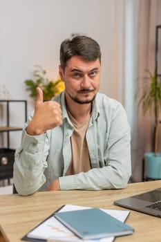 Like. Happy excited Caucasian man looking approvingly at camera showing thumbs up like sign positive something good great news positive feedback. Young guy sitting at home office table. Vertical.