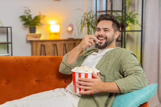 Relaxed Caucasian young man eating popcorn and watching movie sitting on sofa in living room at home. Happy guy with brown hair in casual clothes enjoying suspense film during weekend in apartment.
