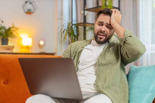Upset young Caucasian young man sitting on sofa at home using laptop having anxiety and stress. Shocked brunette guy reacting to loss, bad news on netbook in living room apartment. Computer virus.