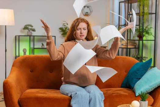 Stressed Caucasian young woman freelancer reviewing business charts and graphs and throwing papers unpaid bills in air sitting on couch in home living room. Angry redhead girl remote working on sofa.