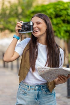 Caucasian young woman tourist traveling on weekends exploring town looking city map, making photo pictures on retro vintage camera outdoors. Happy adult lady traveler on urban city street. Lifestyles.