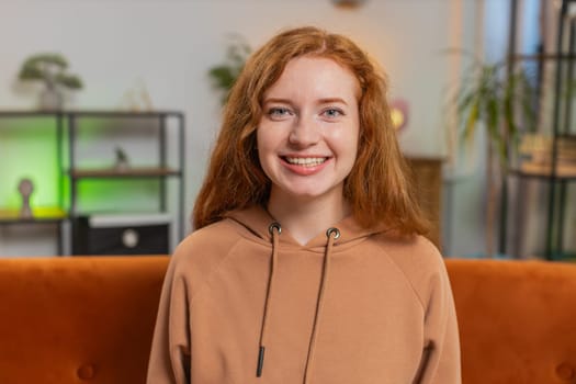 Portrait of smiling young Caucasian woman with long red hair looking at camera. Happy content adult girl in casual clothes relaxing feeling satisfied concept sitting on sofa in living room at home.