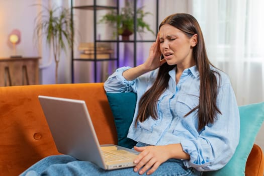 Tired young woman using laptop, suffering from headache problem tension and migraine, stress at home room. Girl working on netbook, sends messages, makes online purchases, watching movies. Lifestyles.