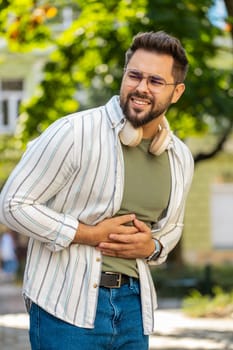 Caucasian young man traveler feeling sudden strong abdominal stomach ache gastritis problem. Upset tourist guy having symptom poisoning diarrhea indigestion peptic ulcer pancreatitis on city street.