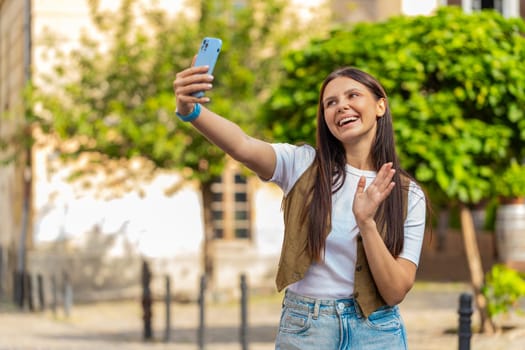Caucasian young woman blogger taking selfie on smartphone, communicating video call online with subscribers, recording stories for social media vlog outdoors. Happy girl standing on urban city street.