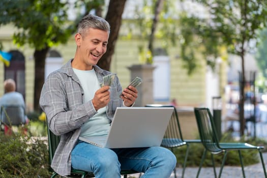 Happy rich man using laptop winner victory counting holding fan of cash money in dollar banknotes sitting on chair city street park. Young male guy tourist planning budget online shopping. Lifestyles
