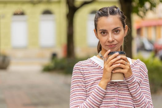 Happy young woman enjoying morning coffee hot drink and smiling outdoors. Relaxing, taking a break. Pretty lovely girl walking through urban city town center street park, drinking coffee to go