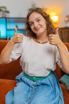 Portrait of excited smiling Caucasian woman looking approvingly at camera showing double thumbs up, like sign, good news, positive feedback. Happy girl sitting on sofa in living room at home. Vertical