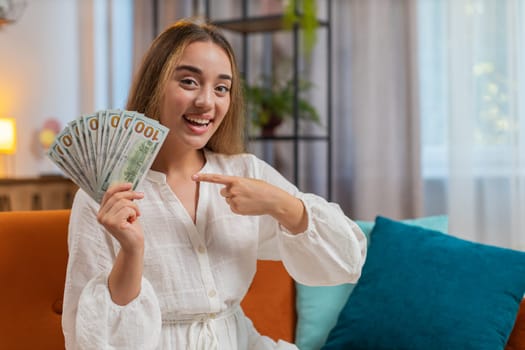 Successful rich Caucasian woman holding waving dollar bill money fan sitting on couch at home room. Happy excited adult girl winning online lottery game planning vacation calculating budget. Lifestyle