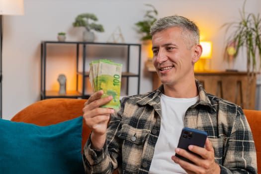 Happy smiling man with gray hair counting money euro cash, use smartphone calculate domestic bills at home indoors. Young guy satisfied of income salary looking at camera sitting in room on couch