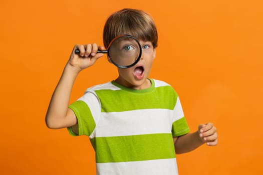 Investigator researcher scientist preteen child boy kid holding magnifying glass near face, looking into camera with big zoomed funny eye, searching, analyzing. Young children on orange background