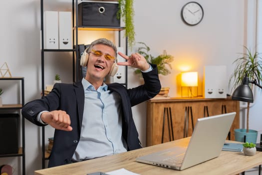 Happy relaxed young businessman working on laptop at office wearing headphones and sunglasses listening favorite energetic disco music, dancing. Freelancer man relaxing, taking a break, celebrating.
