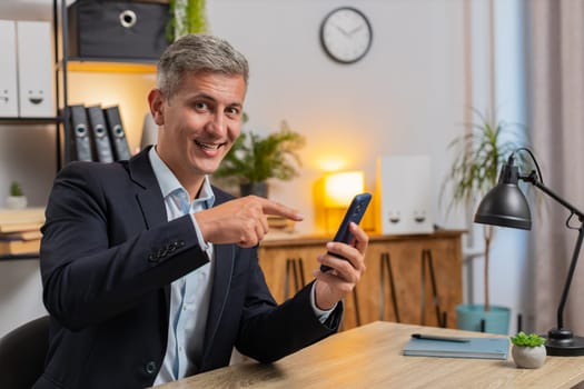 Confident young businessman using smartphone application online pointing showing on mobile screen app, remote distance job working from office. Handsome guy freelancer looking at camera. Advertising