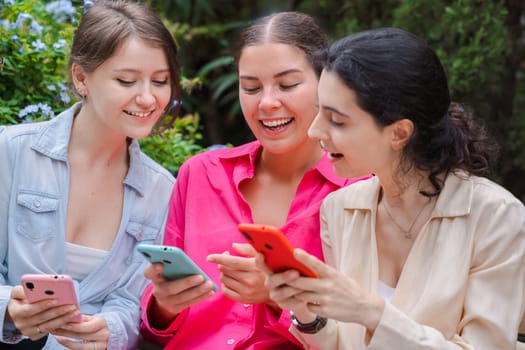 Three young Caucasian women sit on a wooden bench in a green park,smiling while using smartphones.they look happy and engaged,enjoying a casual outdoor moment with modern devices and social media apps