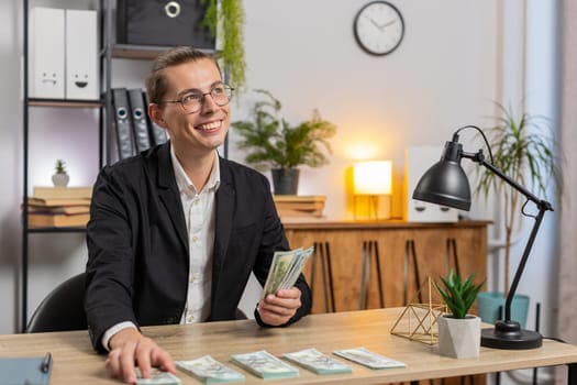 Caucasian successful businessman in formal suit sitting at workplace in home office counting money dollars salary holding stack of dollars, analyzes budget finances. Young freelancer sitting at table.