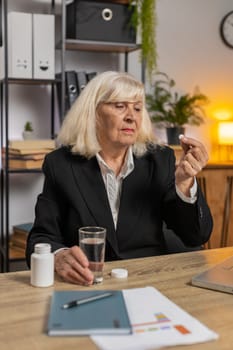 Sick upset senior businesswoman grandmother working on laptop computer suffering from headache at home office. Unhealthy Caucasian freelancer elderly woman taking medicine pill from bottle in room