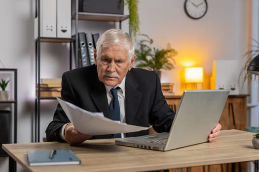 Senior freelancer accountant working with financial graph documents, calculate taxes for charts and reports at home office desk. Businessman grandpa working on laptop computer planning expenses budget