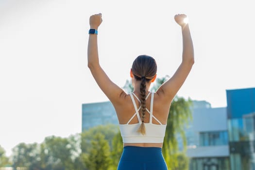Rear view of excited Caucasian athletic woman runner raising arms celebrating success after jogging in urban city park on sunny day. Jogger champion winning marathon. Active sportswoman motivation.
