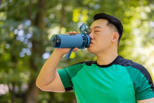Sporty athletic thirsty guy Chinese man runner jogger drinking water after workout in park outdoors drink sport bottle, healthy habit active young Asian lifestyle refreshment hydration looking away.