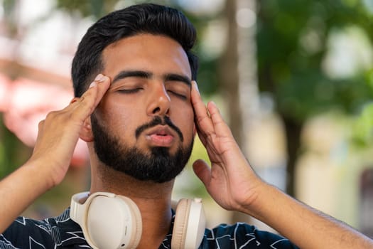 Displeased exhausted Arabian man rubbing temples to cure headache problem suffering from tension migraine stress. Young bearded guy tourist grimacing, pain sitting on chair on city urban street park.