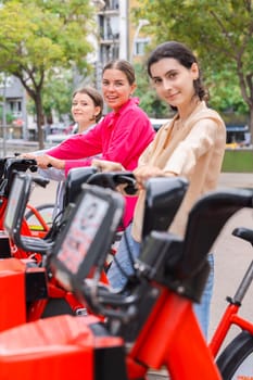 Happy young women renting bicycles in an urban park, smiling and preparing for a fun ride. Dressed casually, they enjoy a lively outdoor setting surrounded by trees and modern city buildings.Vertical