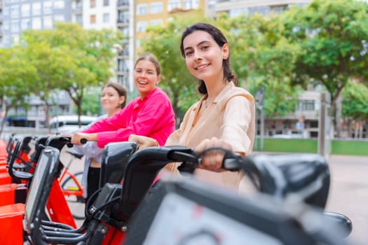 Happy young women renting bicycles in an urban park, smiling and preparing for a fun ride. Dressed casually, they enjoy a lively outdoor setting surrounded by trees and modern city buildings.
