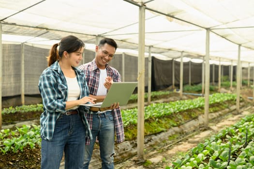 Male and female farmers working together in a greenhouse, using a laptop to monitor crop data.