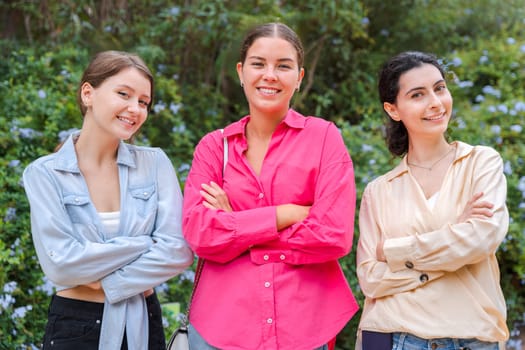 Three young Caucasian women stand outdoors with their arms crossed and a smiling expression. They look confident and determined, posing together in front of the vegetation looking at camera.