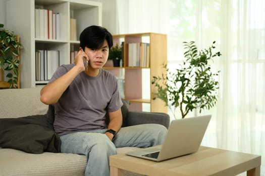 Young asian man talking on smartphone and using laptop in cozy living room.