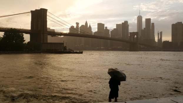 New York City, United States - 11 Sept 2023: Brooklyn Bridge rainy foggy day. Manhattan downtown skyline cityscape, World Trade Center. Man with umbrella, Dumbo Pebble Beach. Autumn river, fall in USA