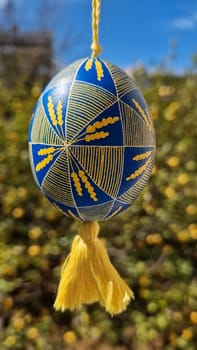 Decorated Ukrainian pysanka in yellow and blue hangs from a string surrounded by lush greenery, embodying traditional Easter symbols and folk artistry during the holiday season.