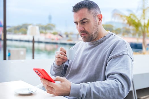 Man with short dark hair and beard drinks coffee while talking on his smartphone.He wears a casual gray sweatshirt and stands outdoors near the water, looking focused in a modern urban setting