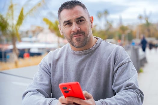 caucasian Man typing on a red smartphone, sitting outdoors by the port with palm trees, wearing a grey sweater, focused on the screen in a relaxed and casual atmosphere.