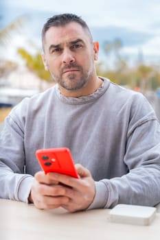 caucasian Man typing on a red smartphone, sitting outdoors by the port with palm trees, wearing a grey sweater, focused on the screen in a relaxed and casual atmosphere. Vertical